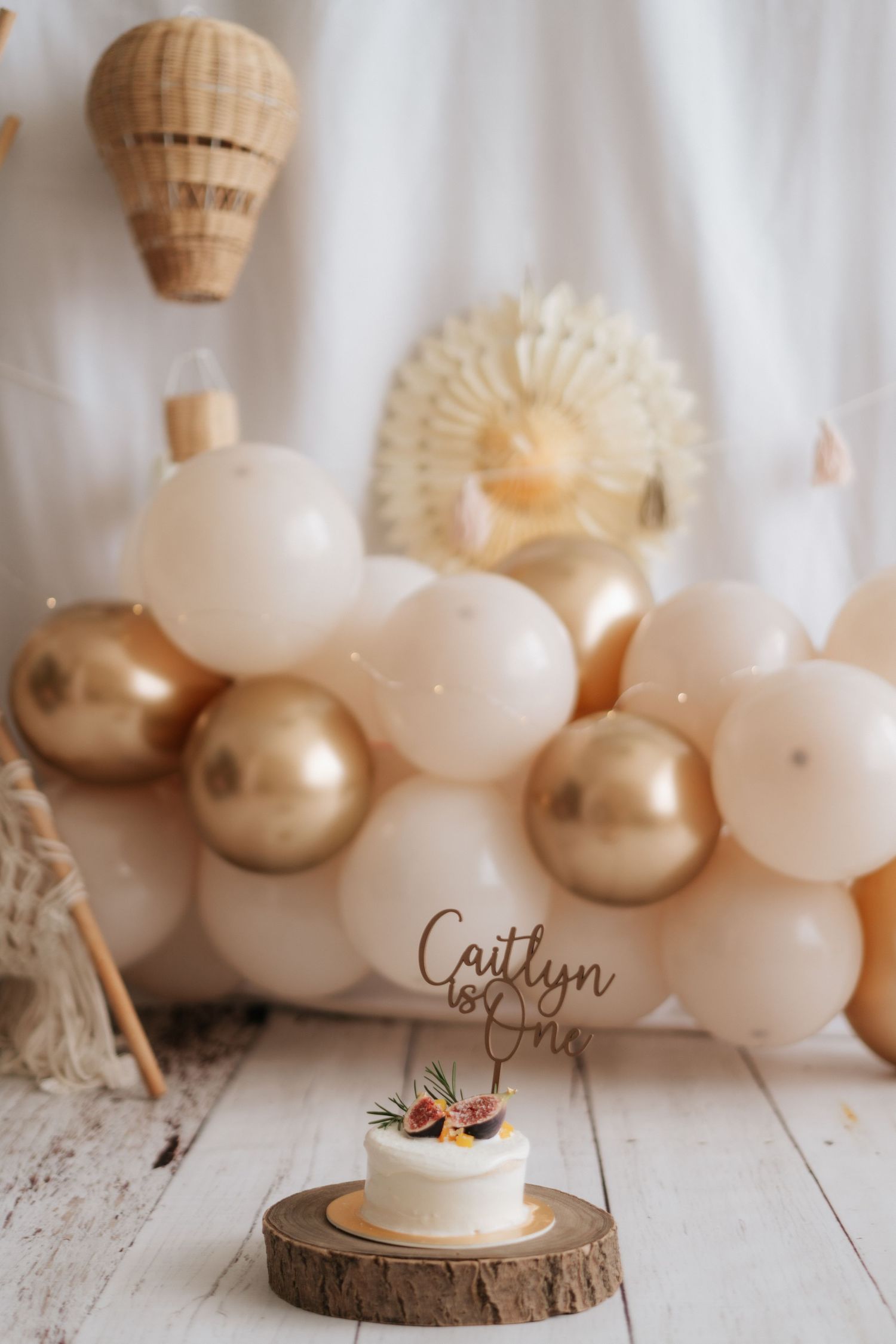 Neutral beige balloon garland with gold accents and wood slices creates an elegant party backdrop on white wooden floor.