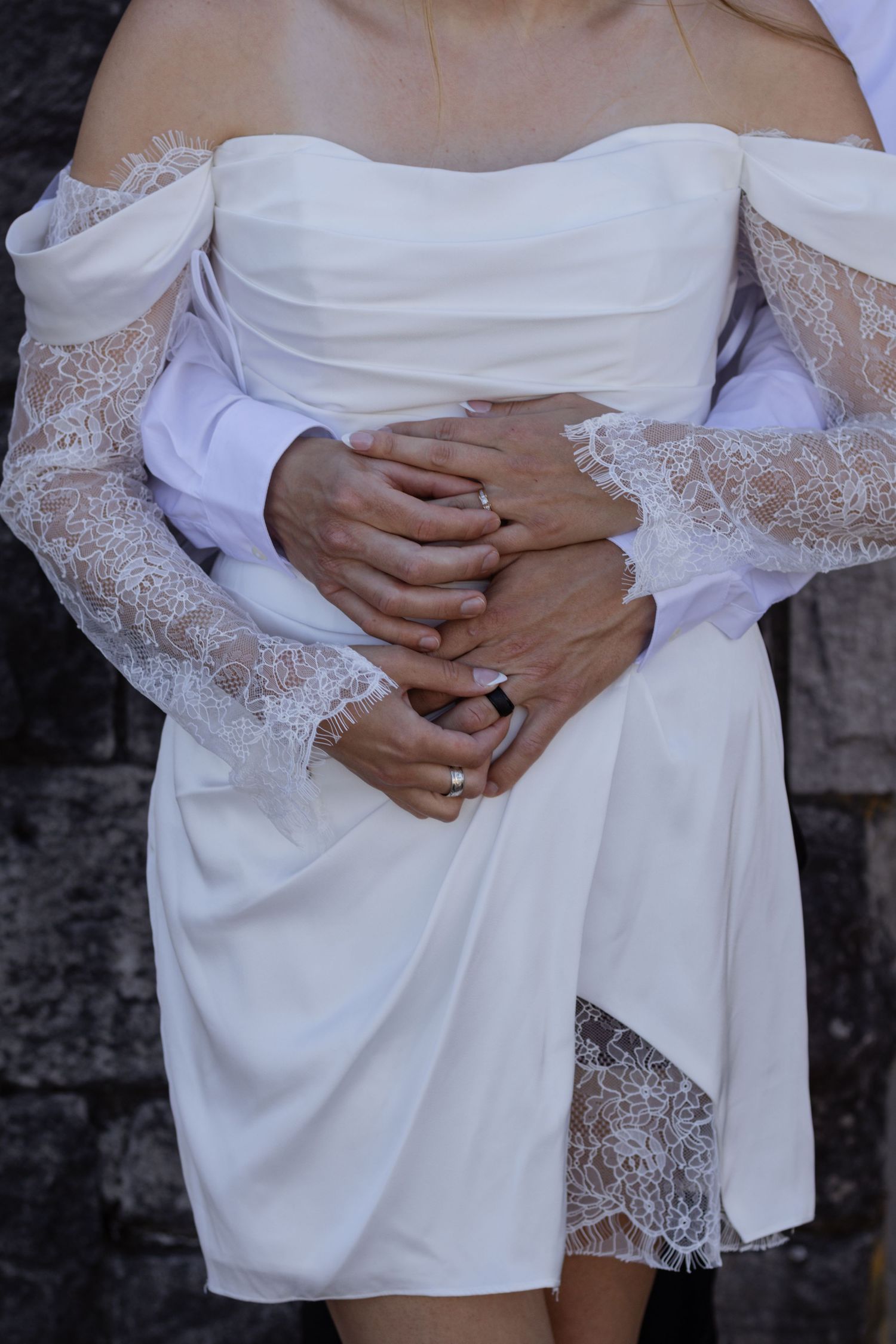 Bride in white off-shoulder lace wedding dress with groom's hands wrapped around her waist, showing wedding ring.