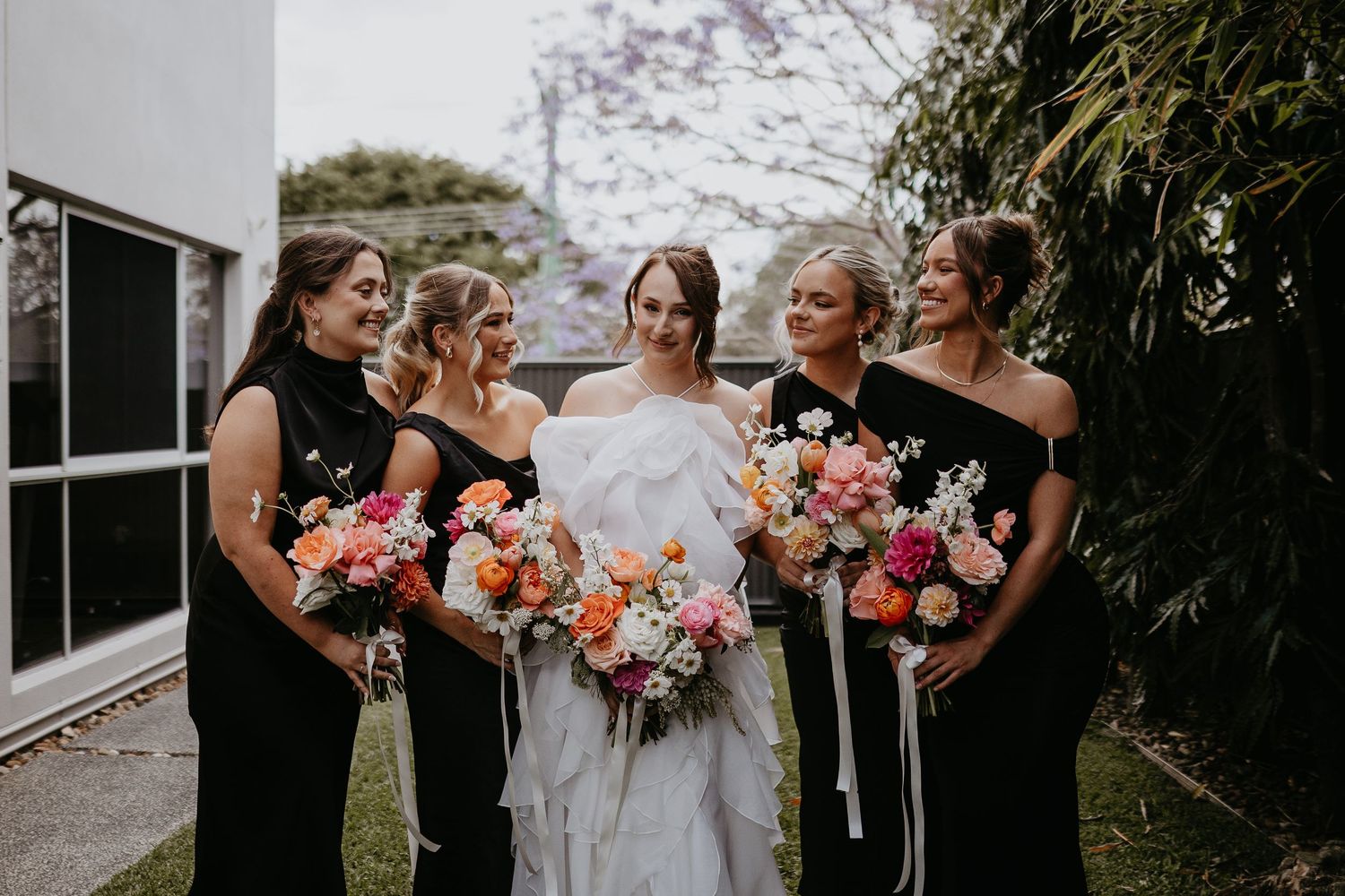 Wedding party members share joyful laughter while holding vibrant pink and white bouquets outdoors.