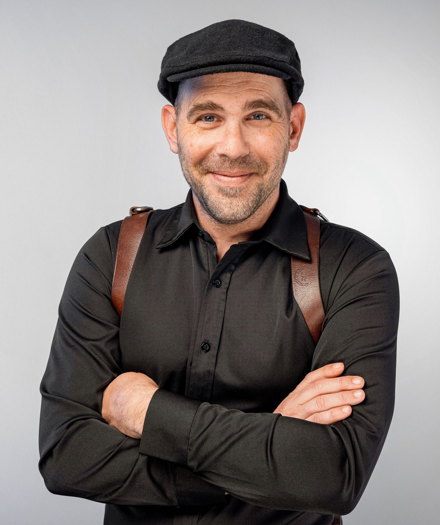 A man in a black shirt and baseball cap poses against a blue backdrop in a professional headshot series.