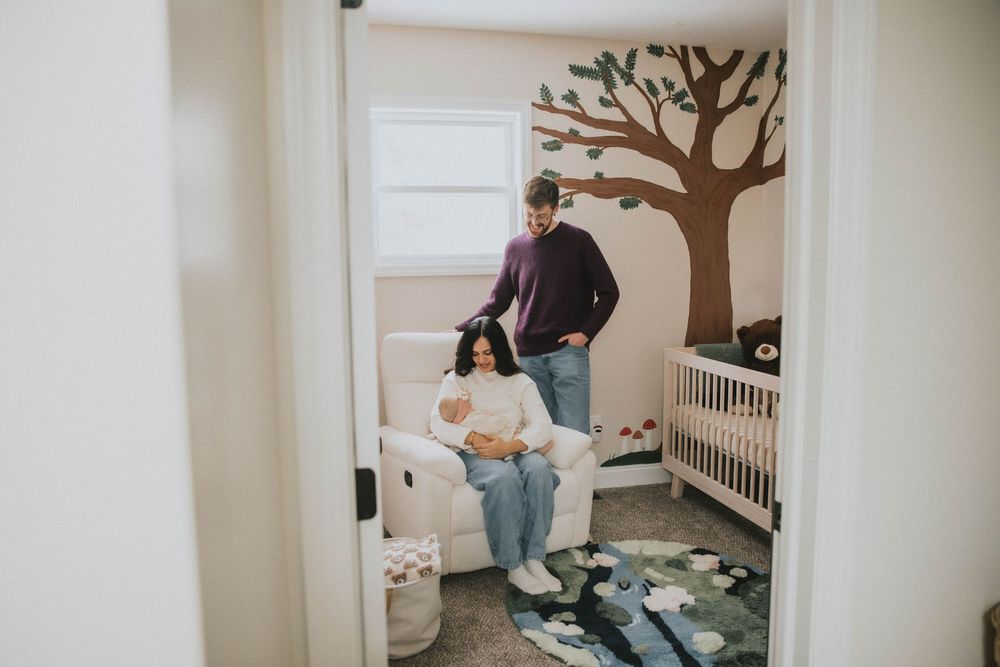 A nursery room with a painted tree mural on the wall, white crib, and two people standing near a rocking chair.