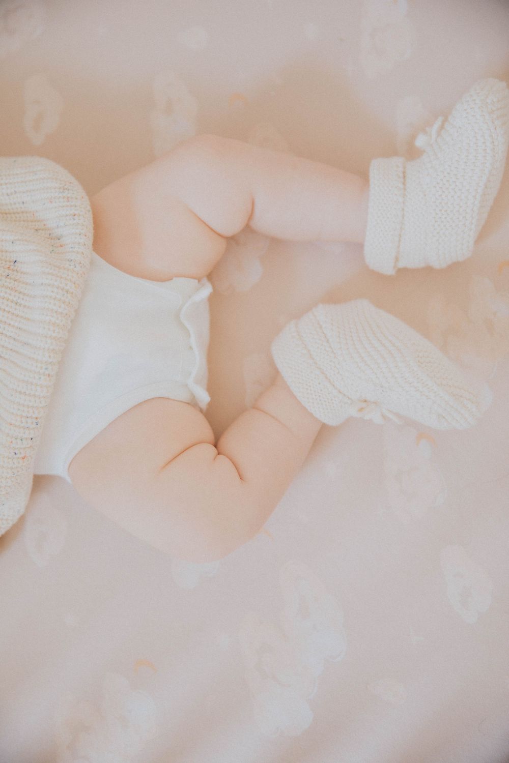A series of soft, dreamy photos showing tiny baby feet wearing white knitted booties against a light beige background.