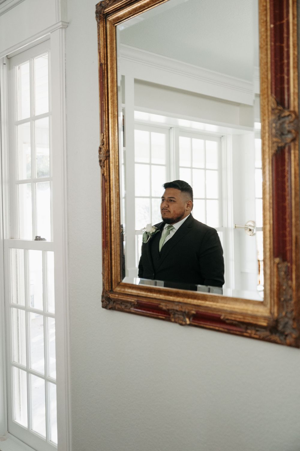 Three identical ornate gold-framed mirrors hang on a white wall next to French windows in an elegant interior space.
