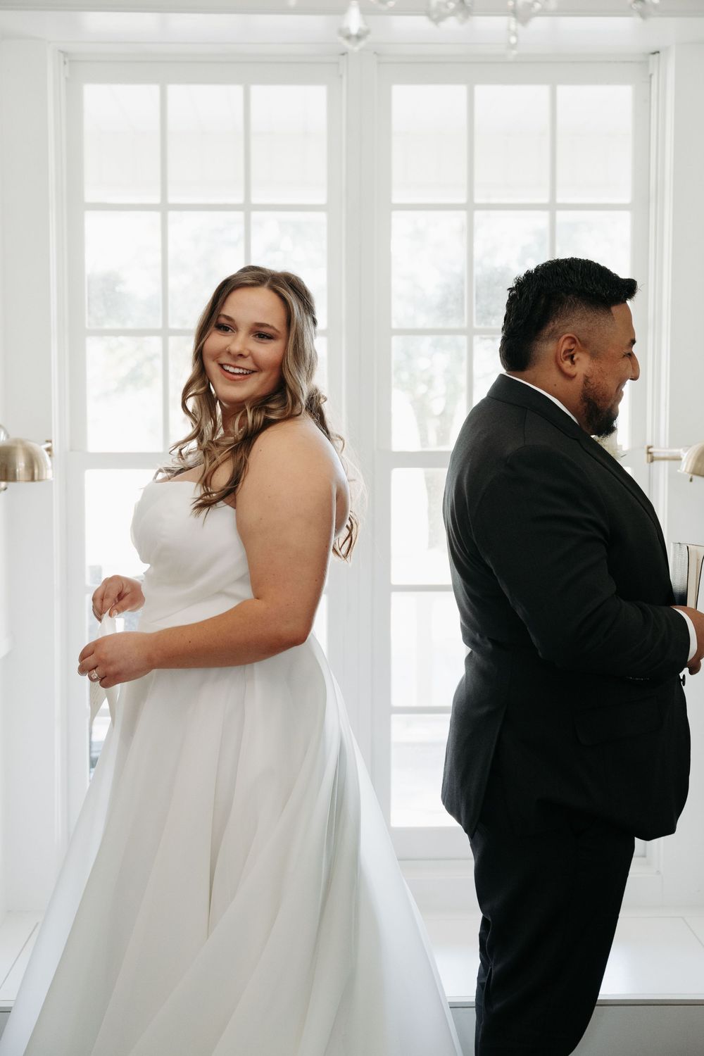 Intimate first look moment between bride and groom in a bright white room with large windows.