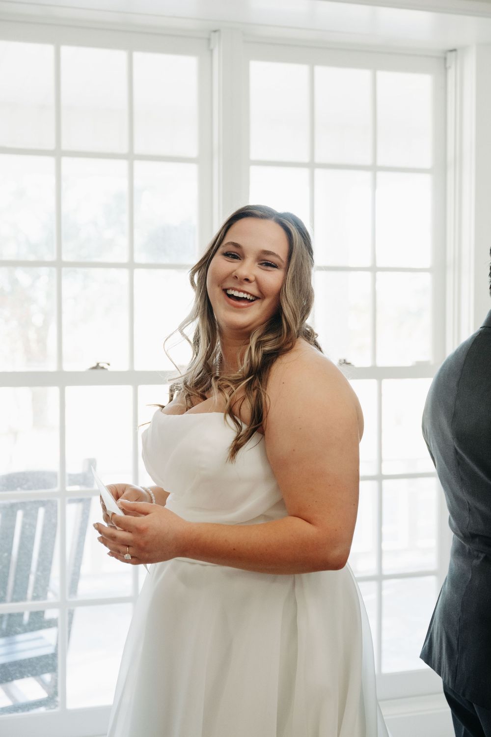 A joyful person in a white wedding dress laughs during a bright indoor celebration by large windows.