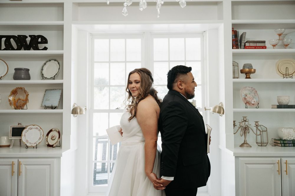 A couple stands back to back in an elegant white room with built-in shelves displaying decorative items and clocks.