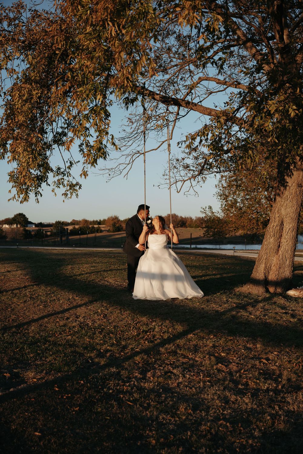 A wedding couple sits beneath autumn trees during golden hour at sunset in a romantic outdoor setting.