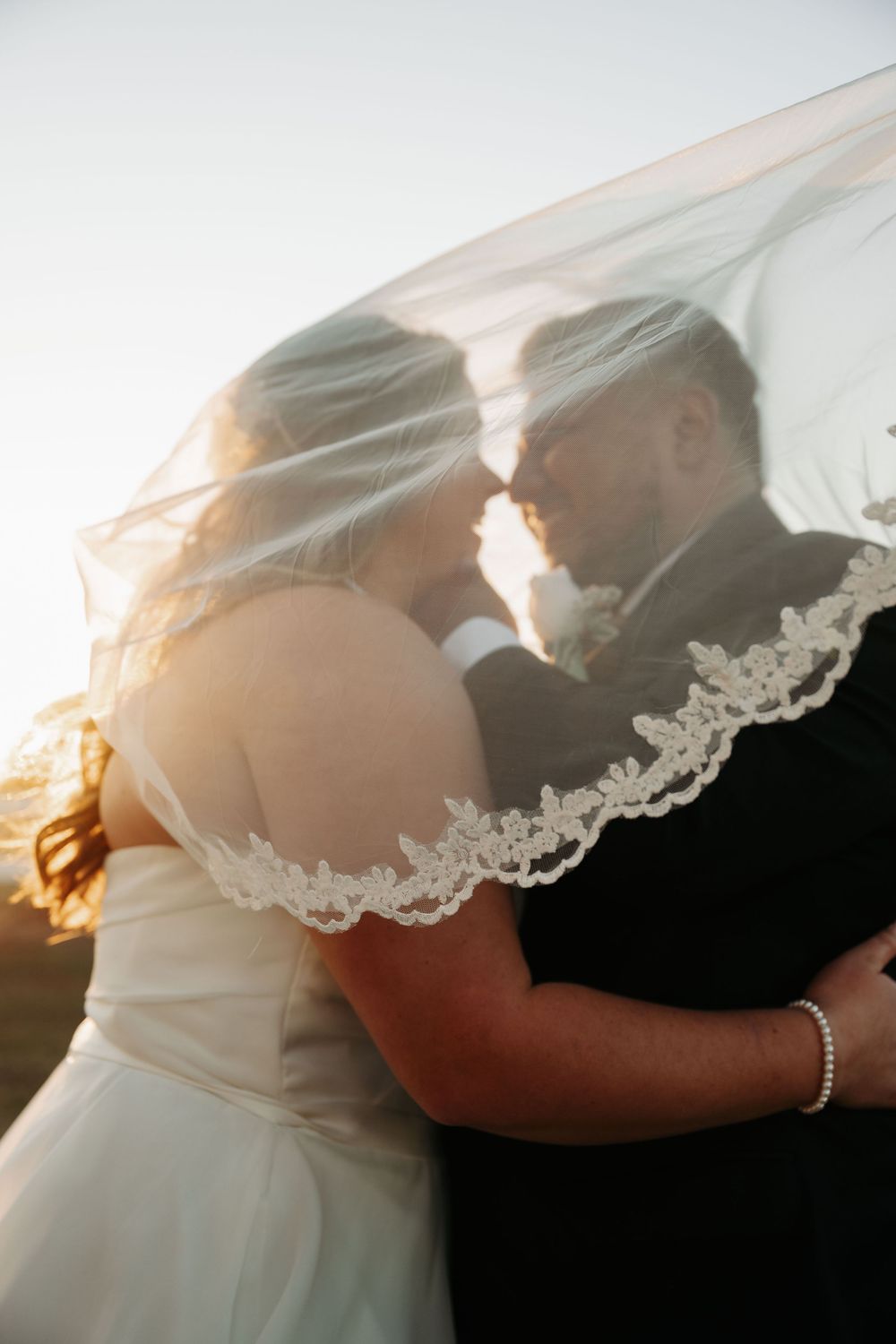Romantic silhouette of newlyweds embracing under delicate lace veil during golden hour.