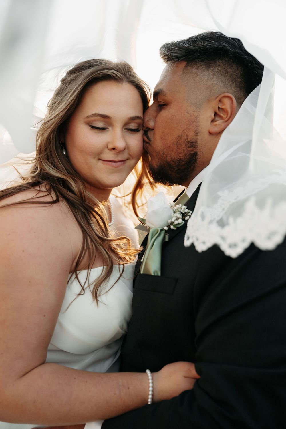 Intimate wedding portrait showing tender moment between newlyweds with flowing veil and white rose boutonniere at sunset.
