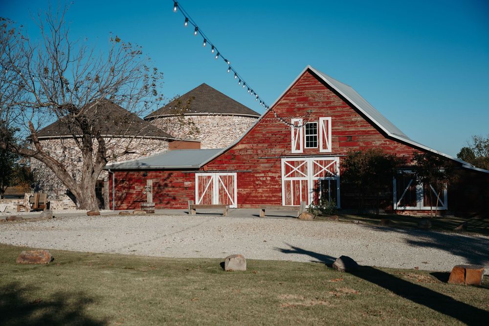 A rustic red barn wedding venue with white trim and a silo stands against a bright blue sky on a sunny day.