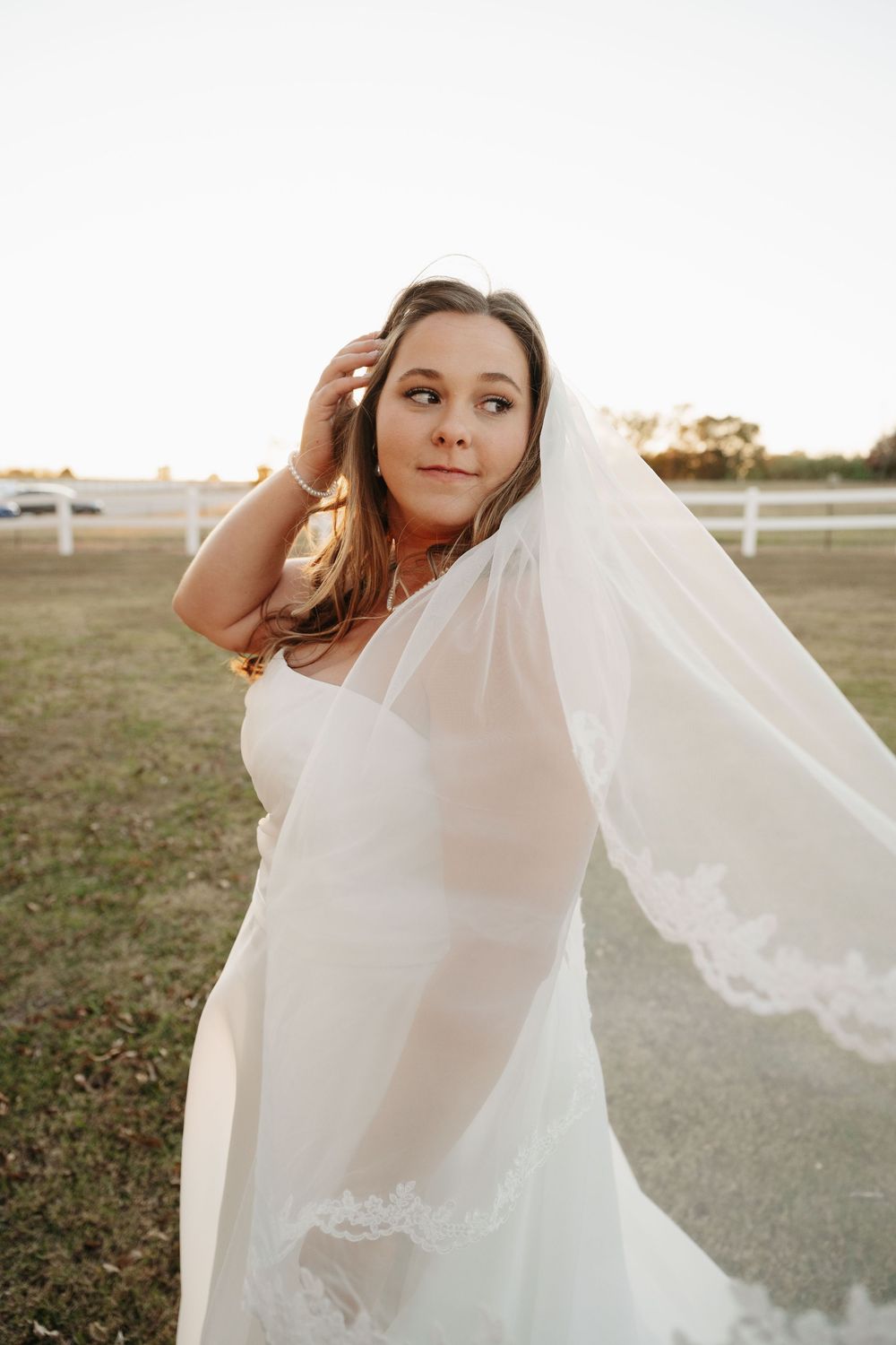 Bride in white dress with flowing veil poses gracefully in an outdoor field at sunset.