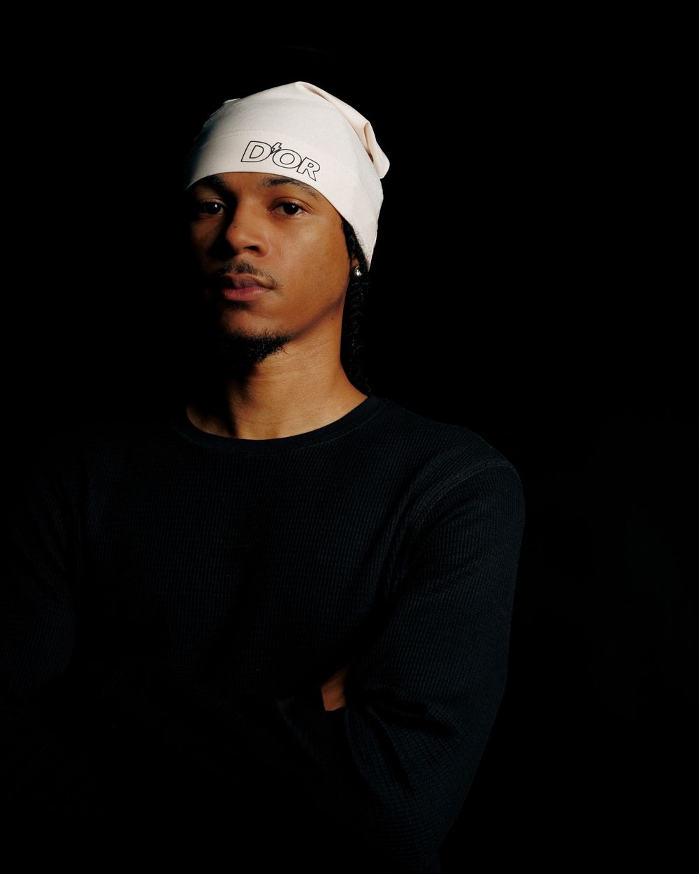 Artistic portrait in dark lighting showing figure wearing white beanie against black background.
