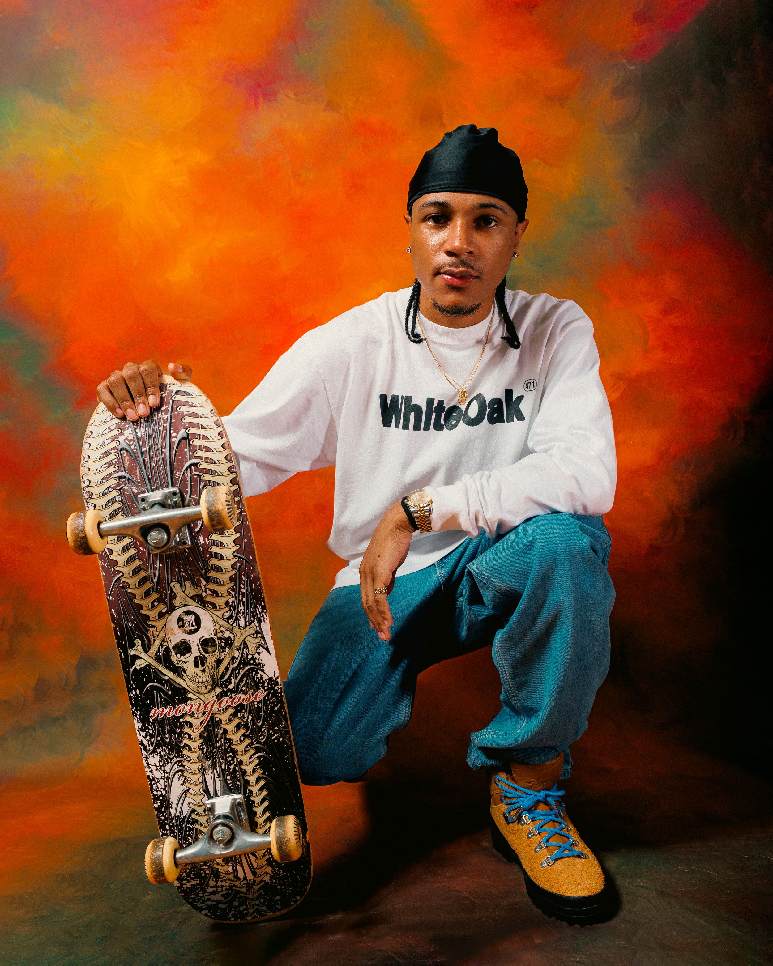 A skateboarder in casual attire poses with their skateboard against a dramatic orange and black backdrop.
