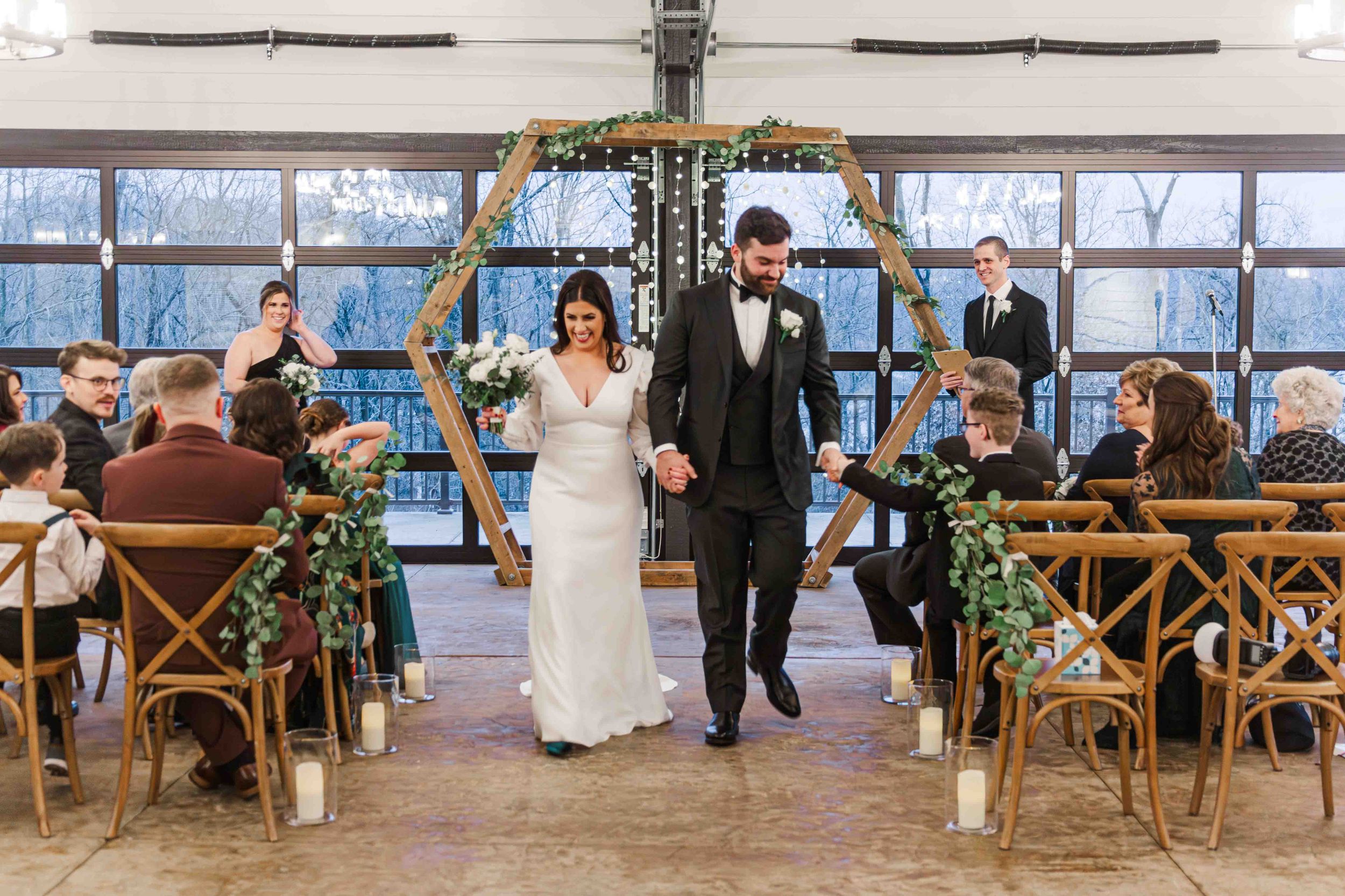 A couple walks down the aisle together after their indoor ceremony with rustic chairs and blue light streaming through windows.
