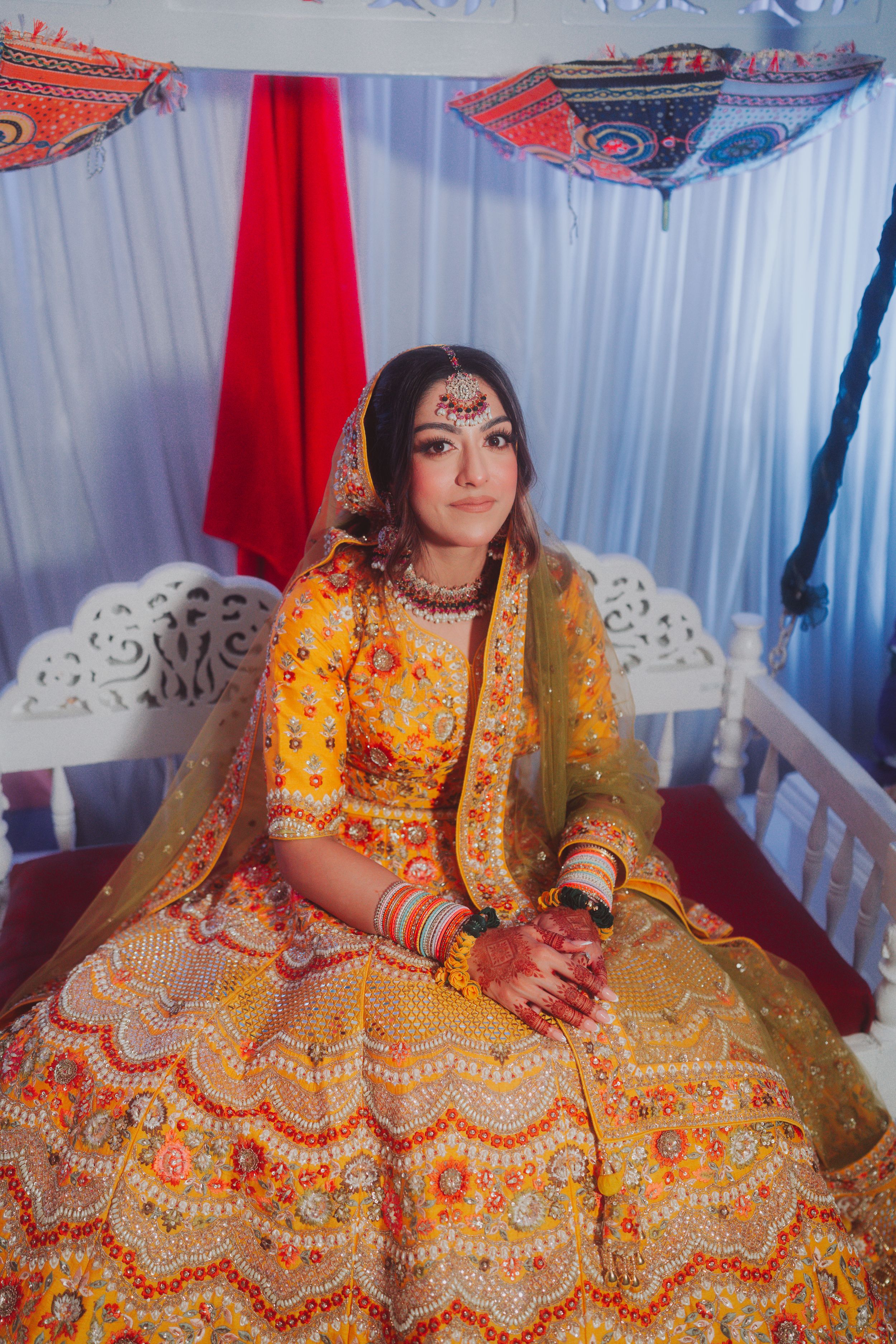 Traditional Indian bride wearing vibrant yellow and orange lehenga with intricate embroidery sits on decorative white chair.