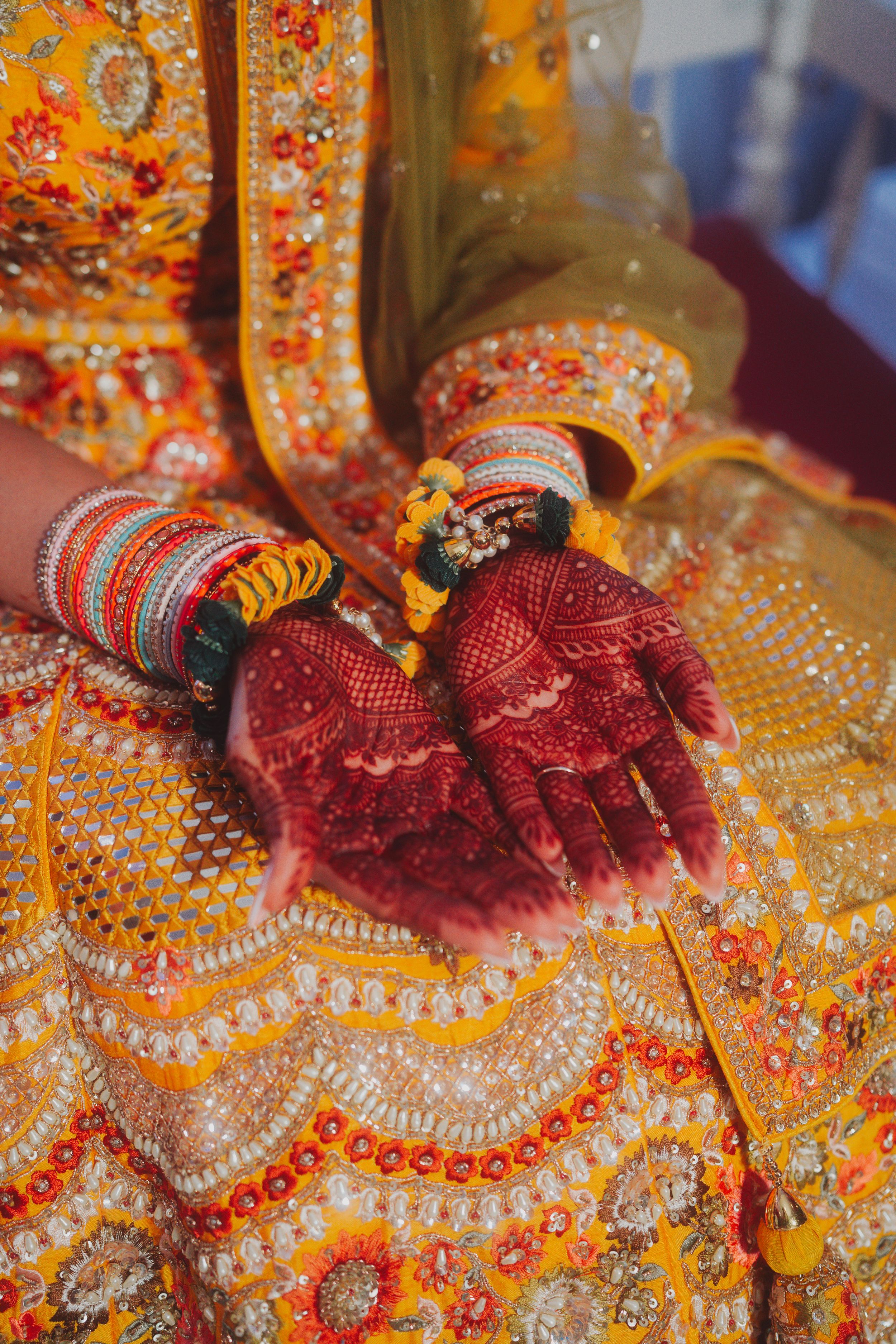 Close-up of bridal hands with henna and red bangles against ornate gold embroidered fabric.