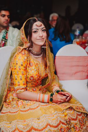 A bride in a vibrant yellow and orange lehenga with traditional jewelry sits at her wedding celebration.