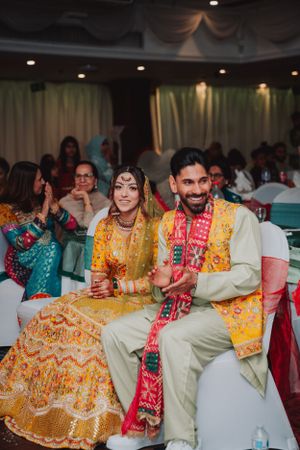 A couple sits together at their wedding celebration wearing traditional yellow and cream outfits.