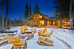 Snow-covered log cabin with Adirondack chairs and fire pit at twilight.