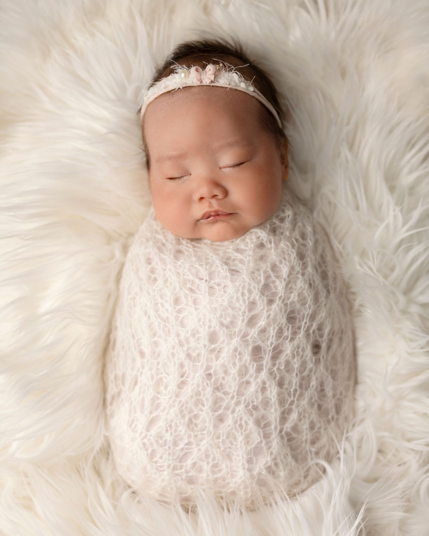 A sleeping newborn wrapped in delicate white knitted lace swaddle rests on soft white fur with a lace headband.