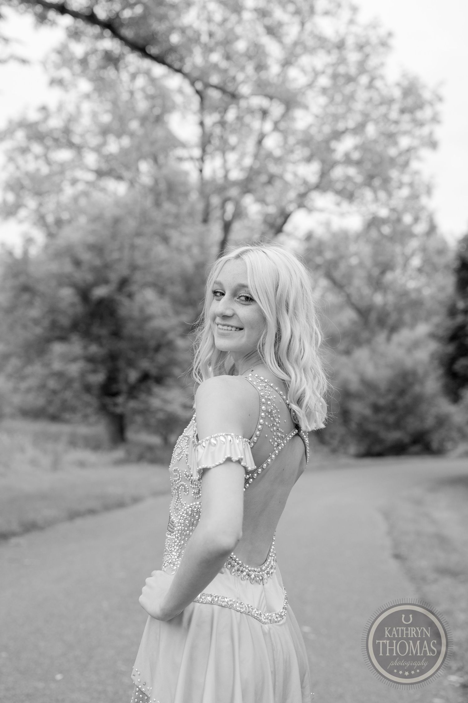 Black and white portrait of a pageant winner in an embellished dress on a scenic pathway.