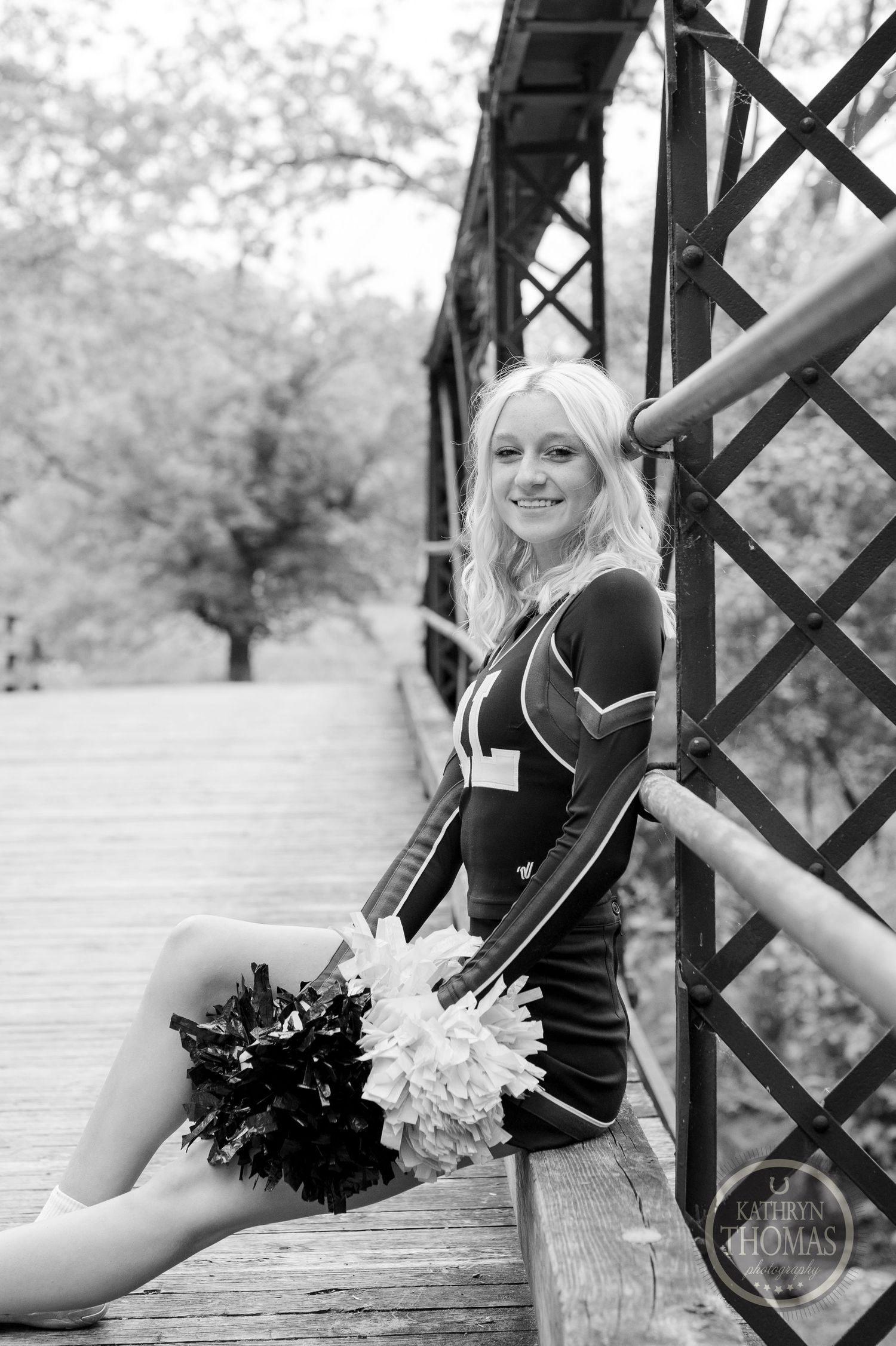 Black and white portrait of cheerleader posing with pom poms on vintage wooden bridge.