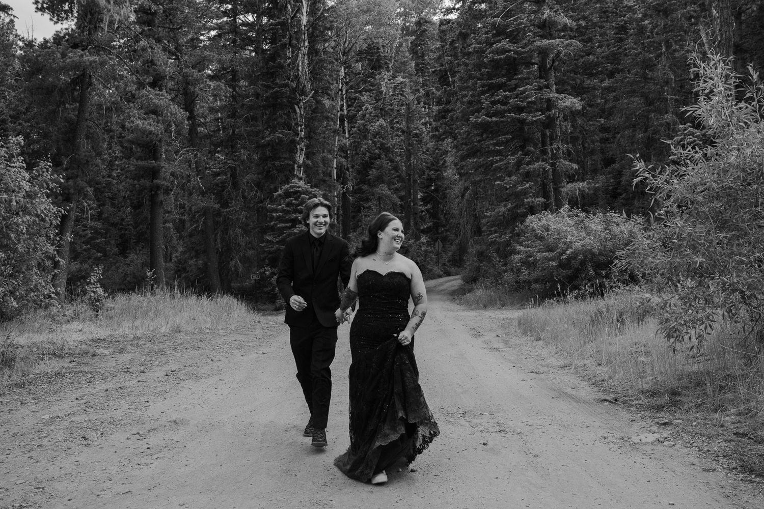 Black and white photo of a couple laughing and running together down a forest dirt road.
