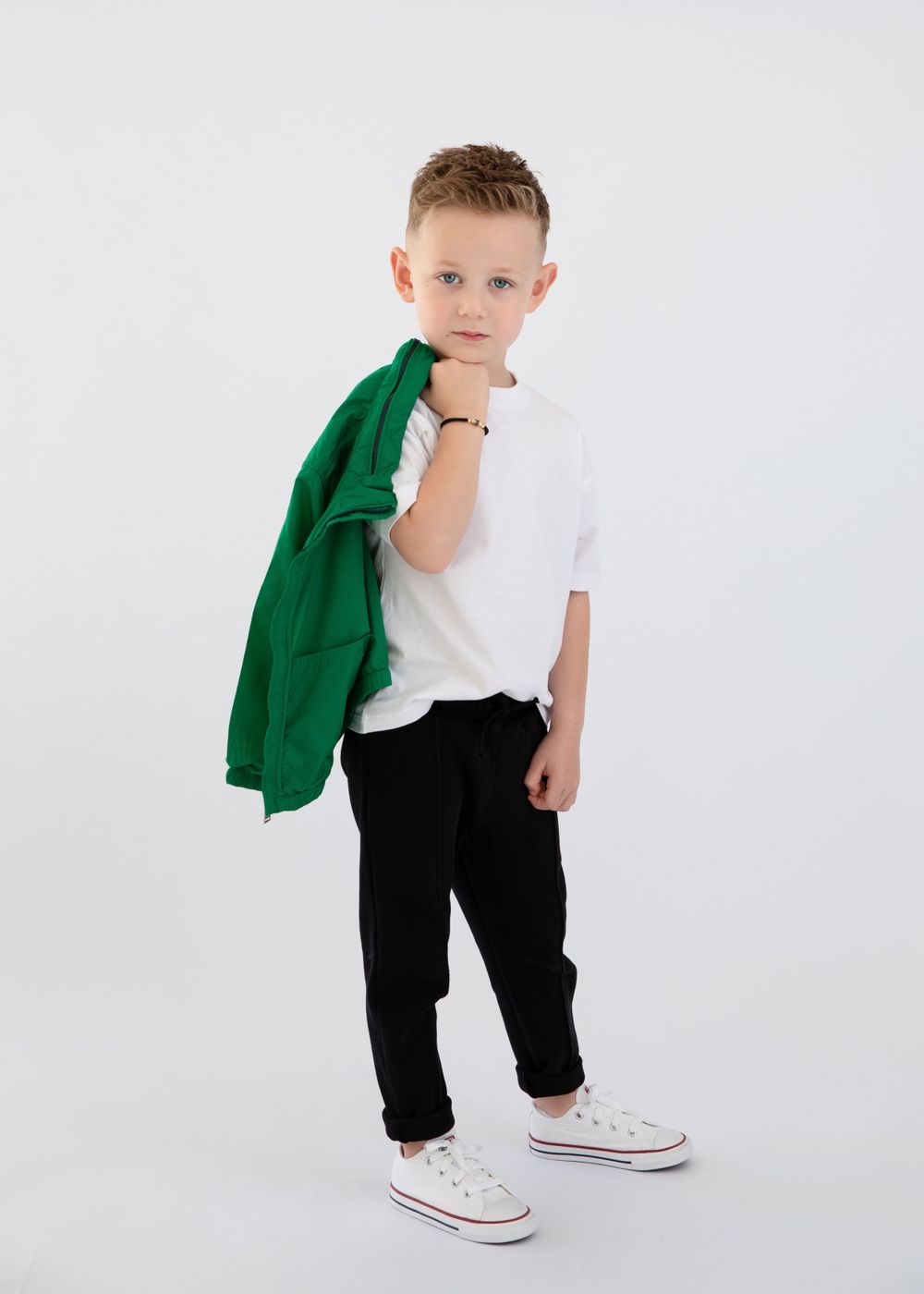Model in casual wear holding green jacket poses with white sneakers against studio background.