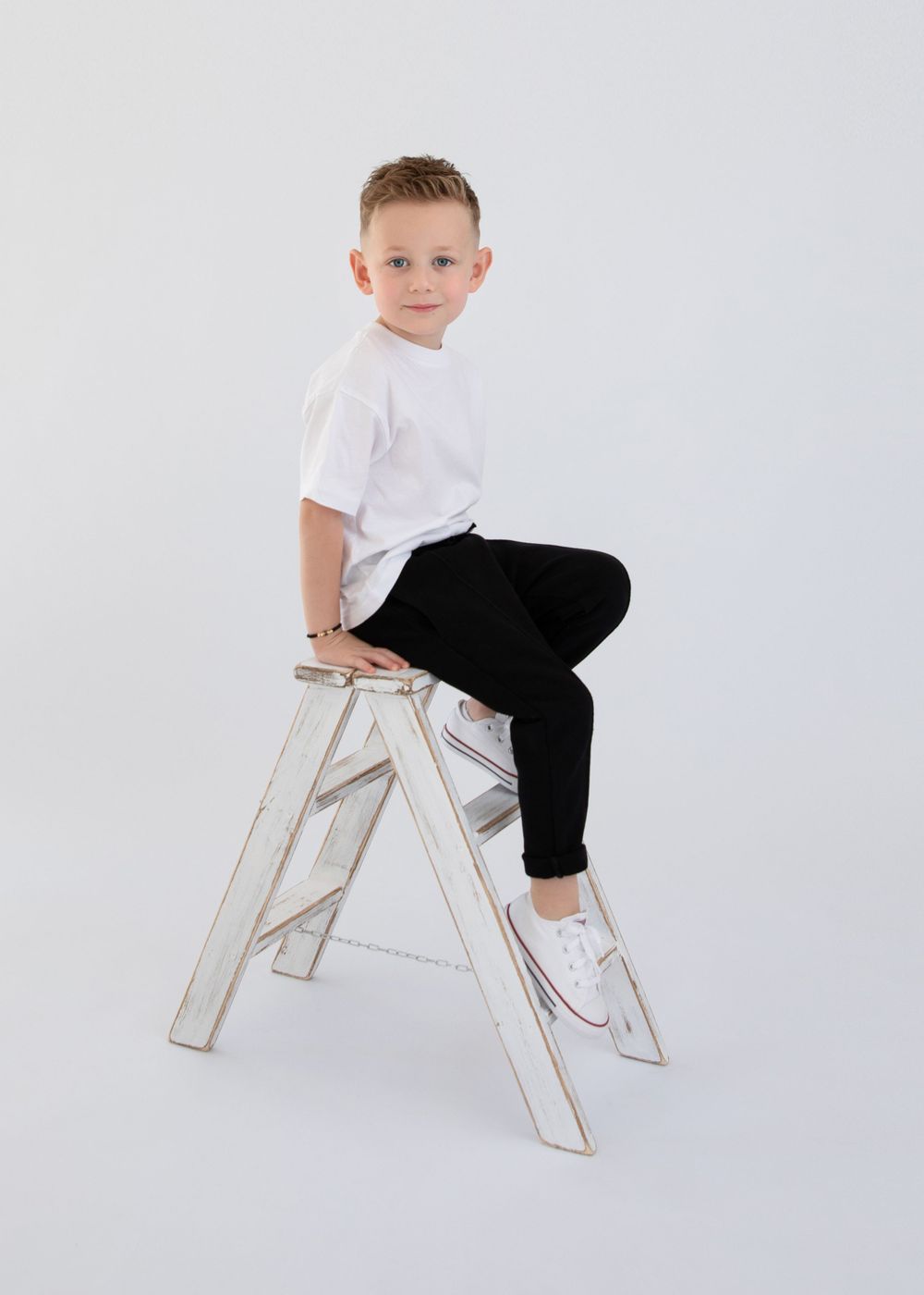 Model in white shirt and black pants sitting on vintage white ladder against studio backdrop.
