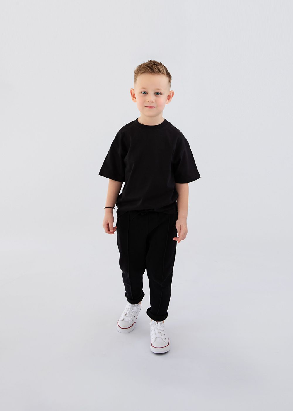 Model wearing all black outfit with white sneakers poses against minimalist background.