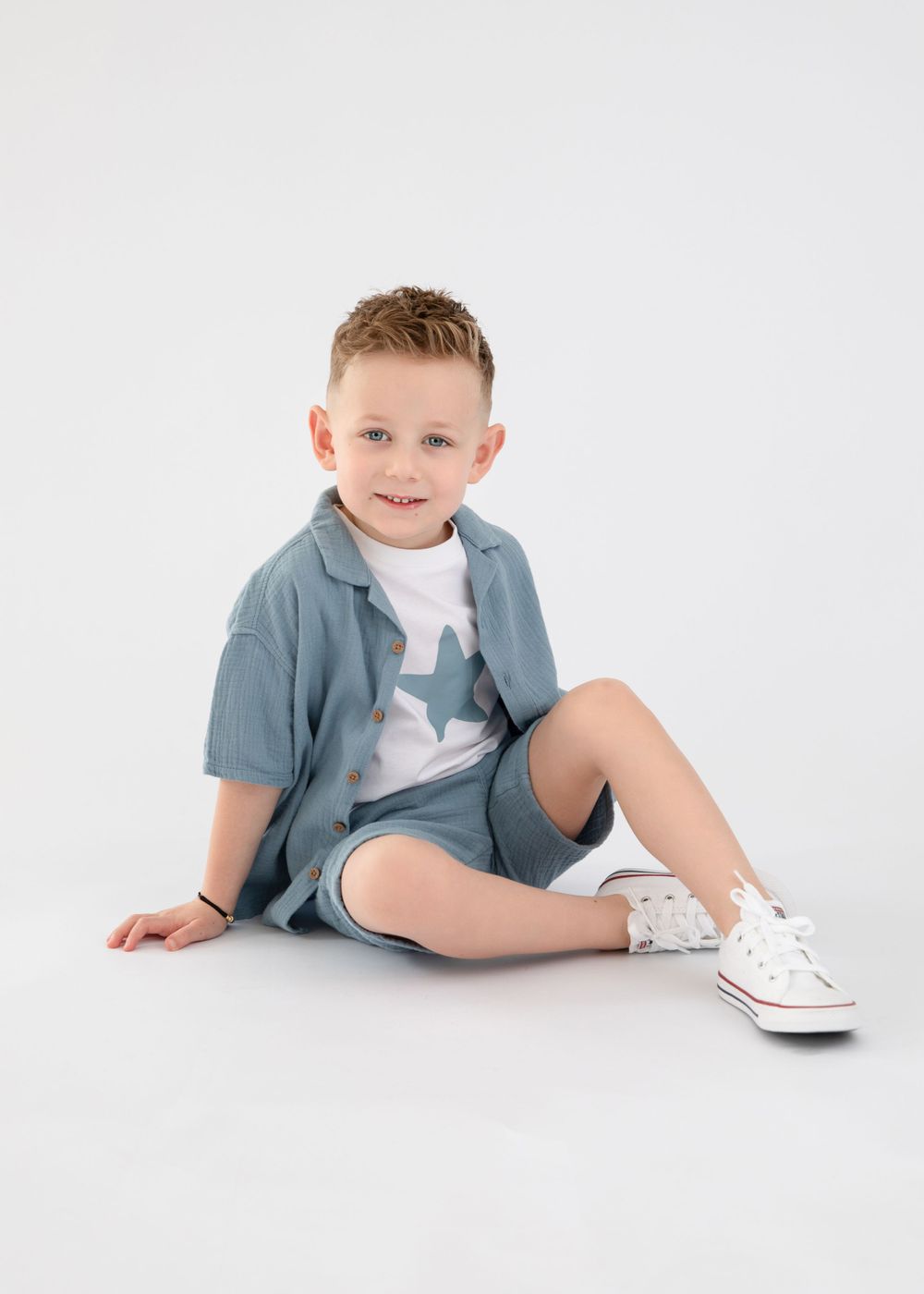 A child in casual blue attire sits on the ground wearing white sneakers against a white background.