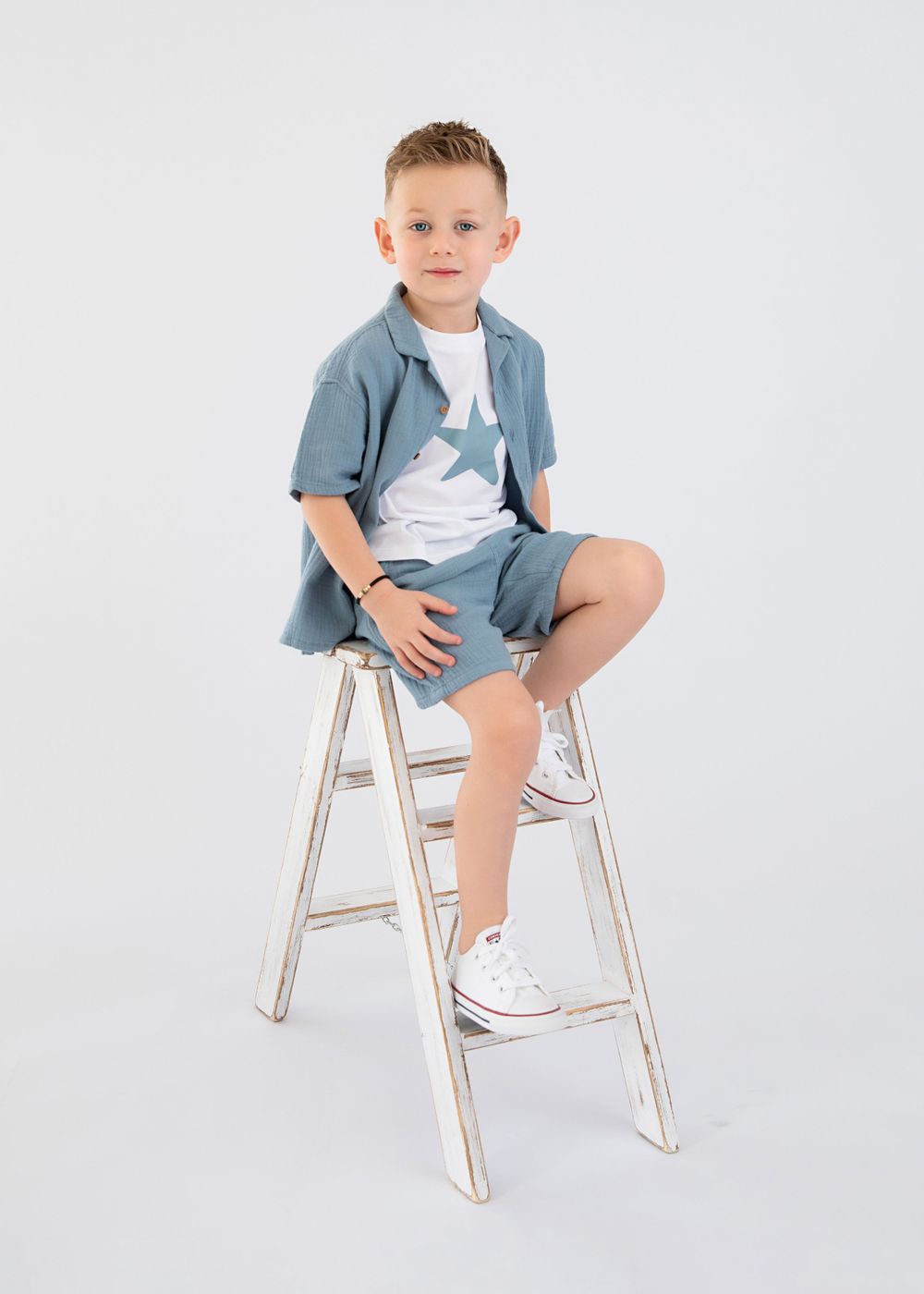 Young model wearing blue shirt and shorts poses on a white ladder stool against light background.