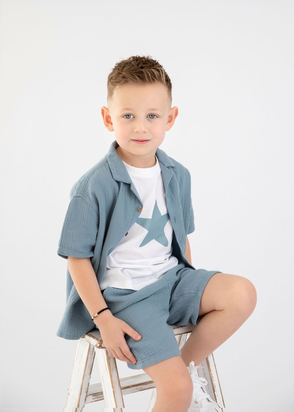Model wearing casual blue outfit sits on white ladder stool during studio photoshoot.