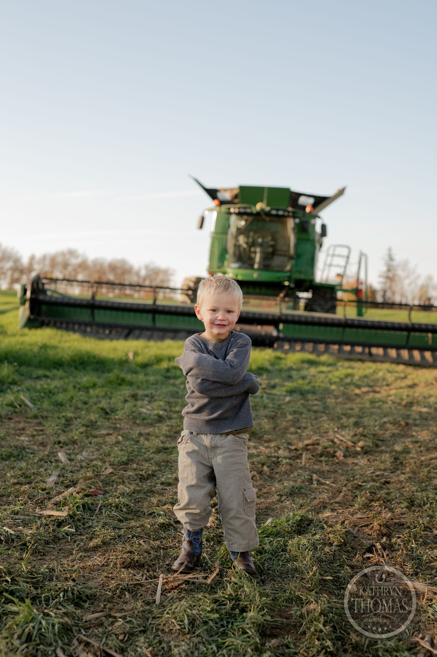 A small child stands confidently in front of a large green combine harvester in a field.