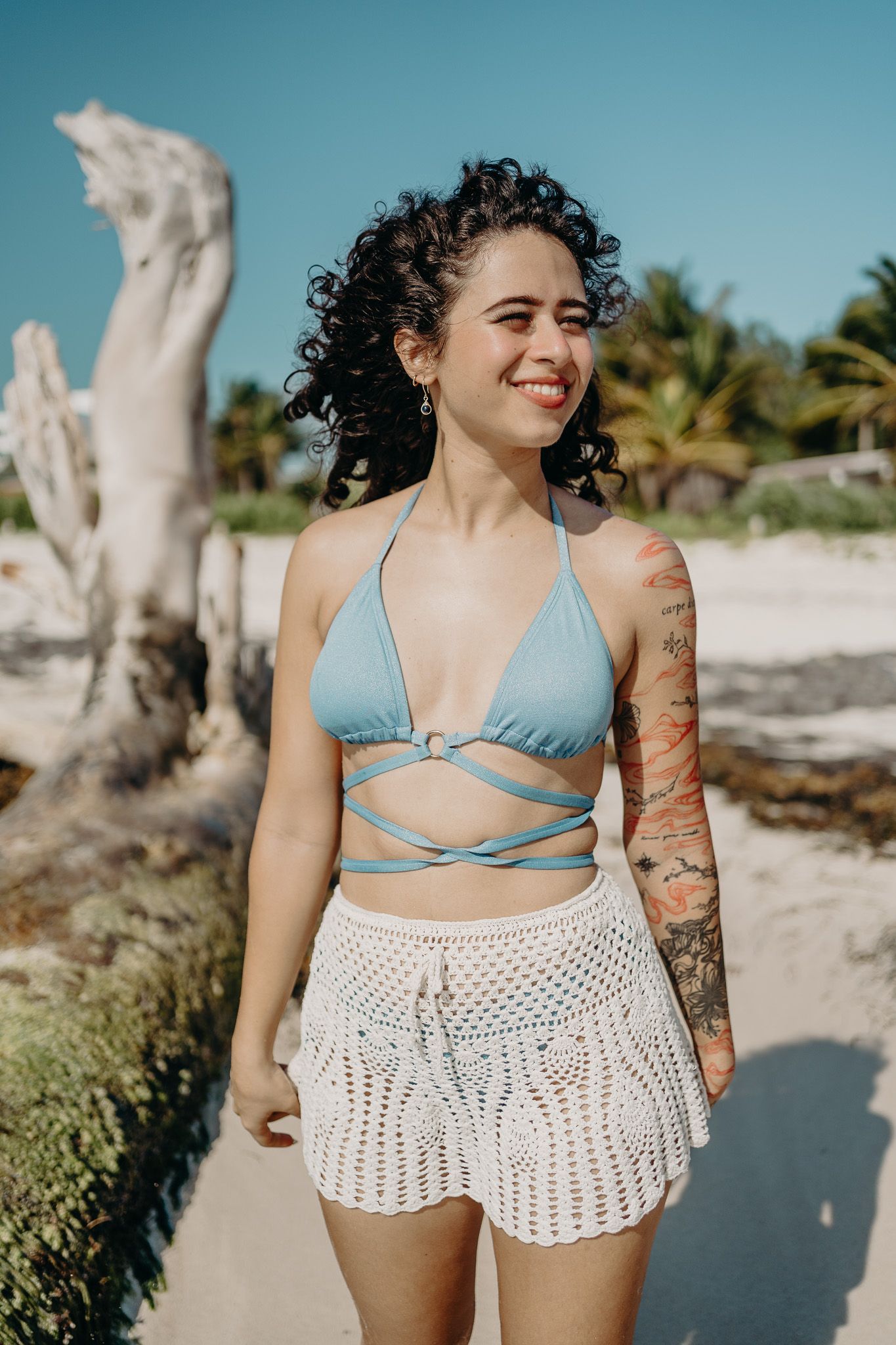 Woman in blue bikini top and crochet shorts standing on beach with driftwood and palm trees photoshoot in Puerto Morelos.