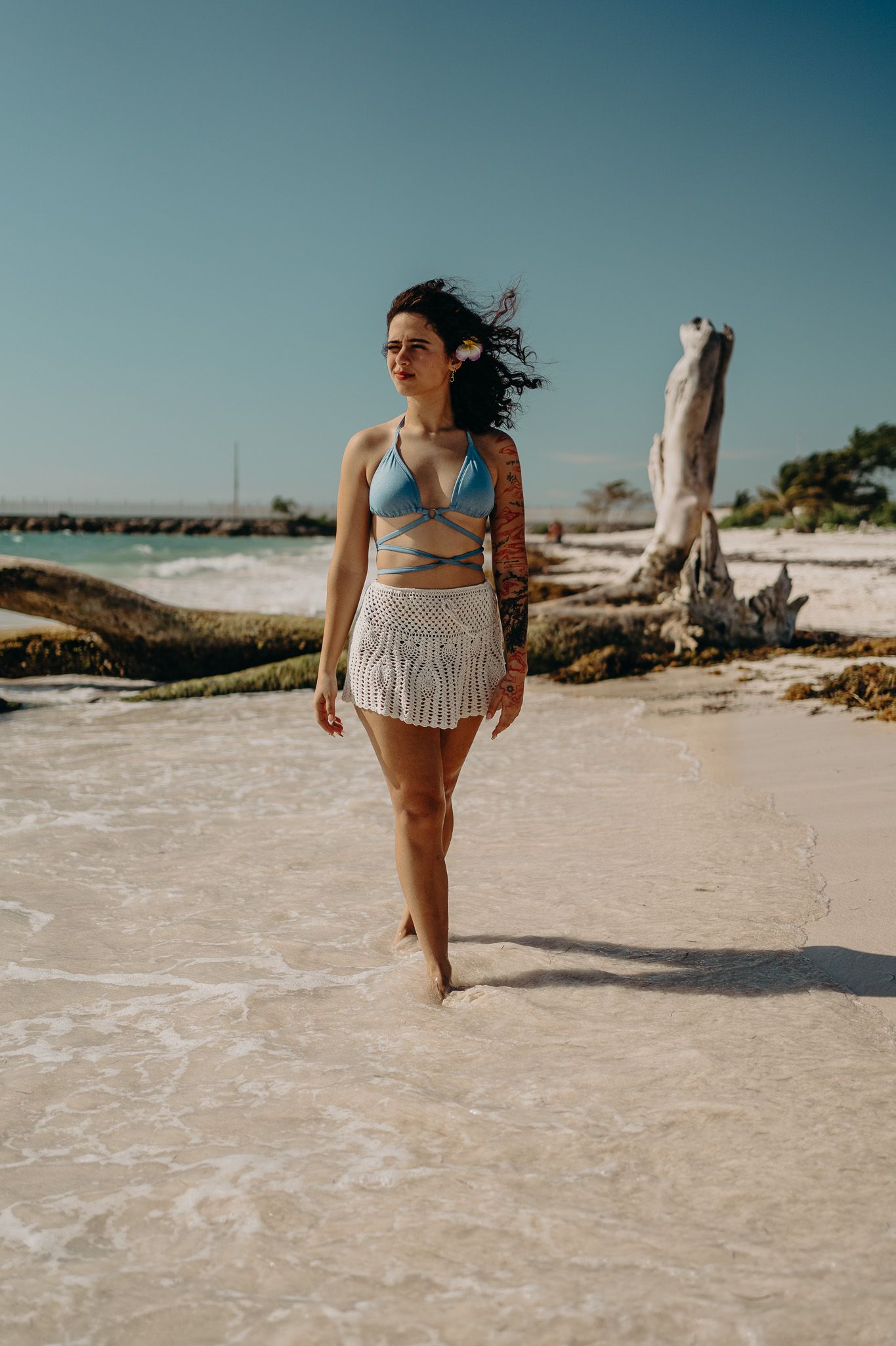 Woman in blue swimwear and white crochet skirt walking along shoreline with driftwood in background photoshoot in Puerto Morelos.