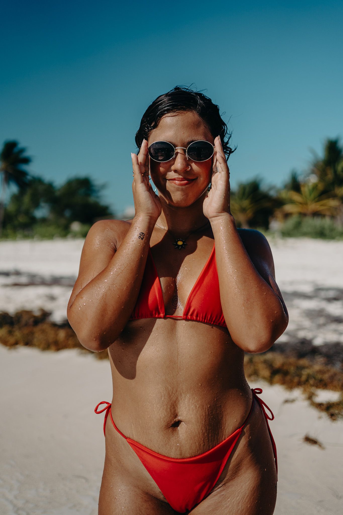 Person in red swimwear posing playfully with sunglasses on tropical beach photoshoot in Puerto Morelos.
