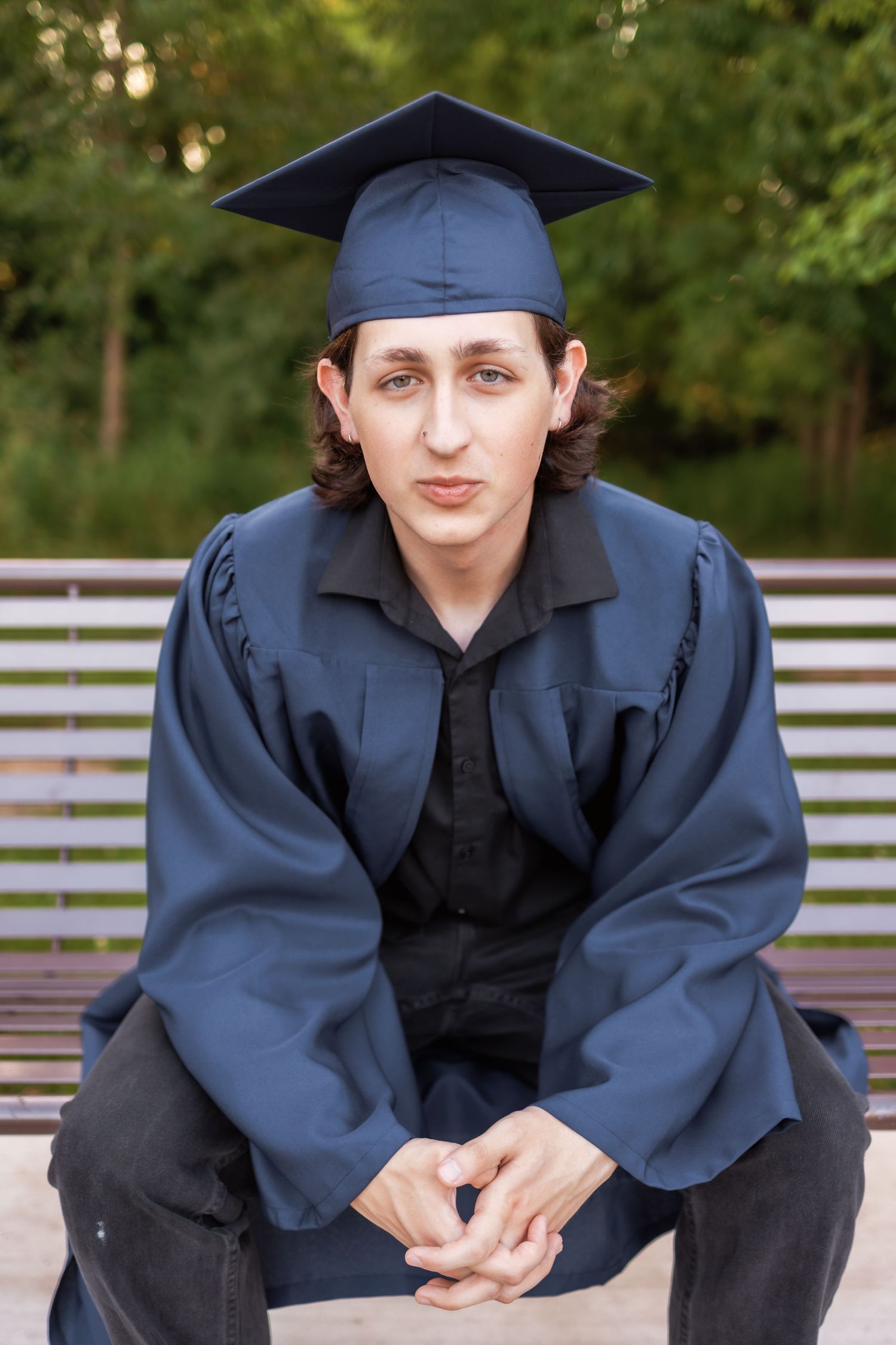 A person wearing dark blue graduation cap and gown sits on a bench outdoors.