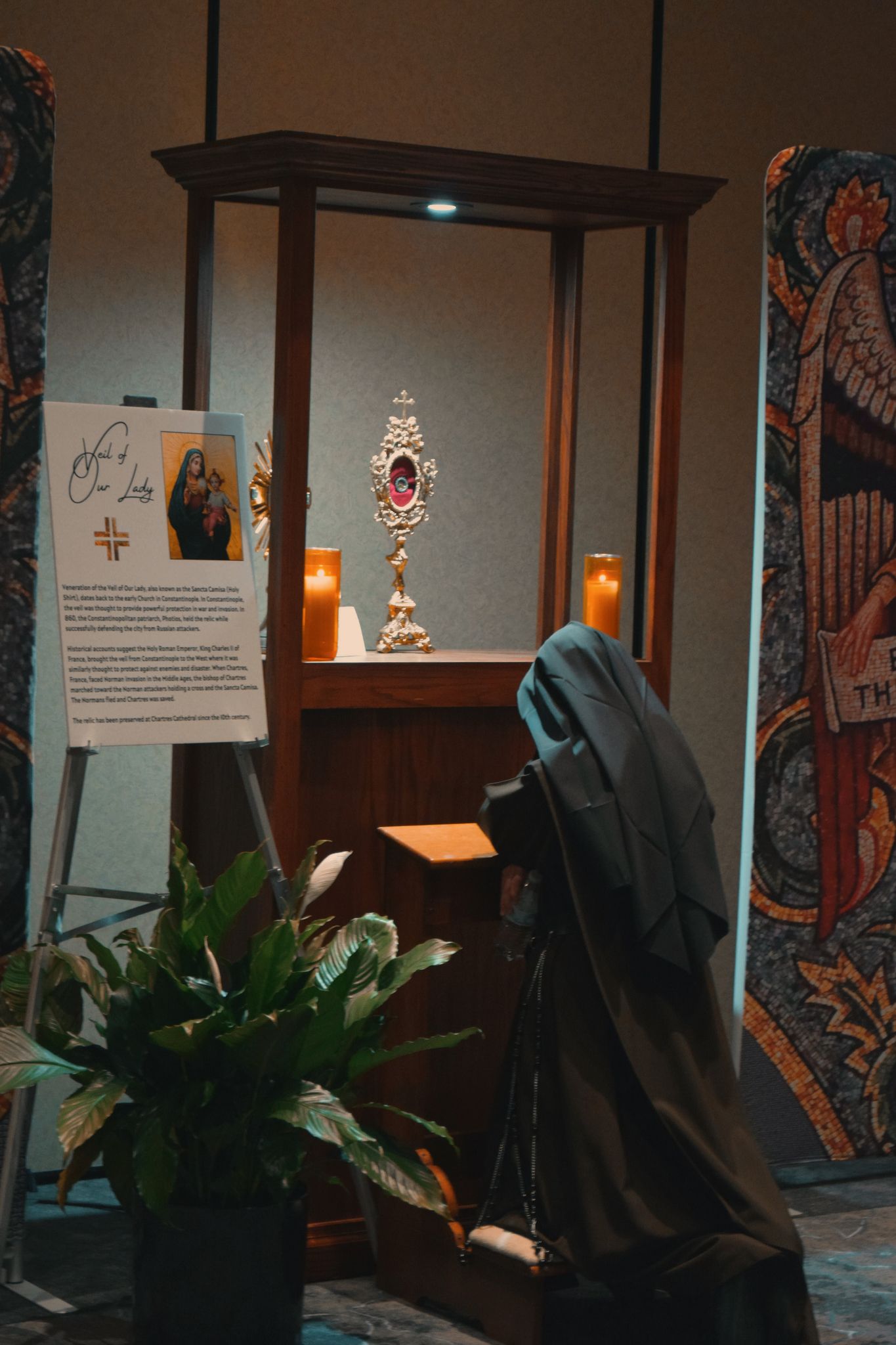 A religious display featuring an ornate reliquary in a wooden case with a hooded figure viewing it and decorative tapestries nearby.