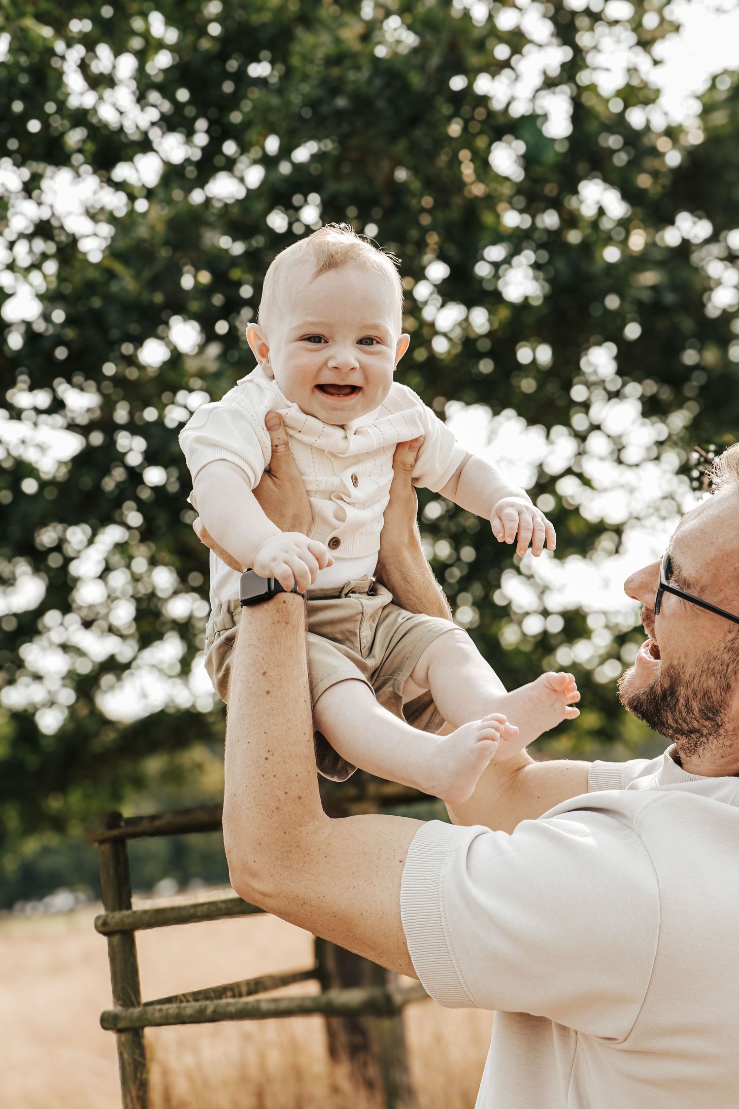 Happy baby in beige shorts being lifted up outdoors.