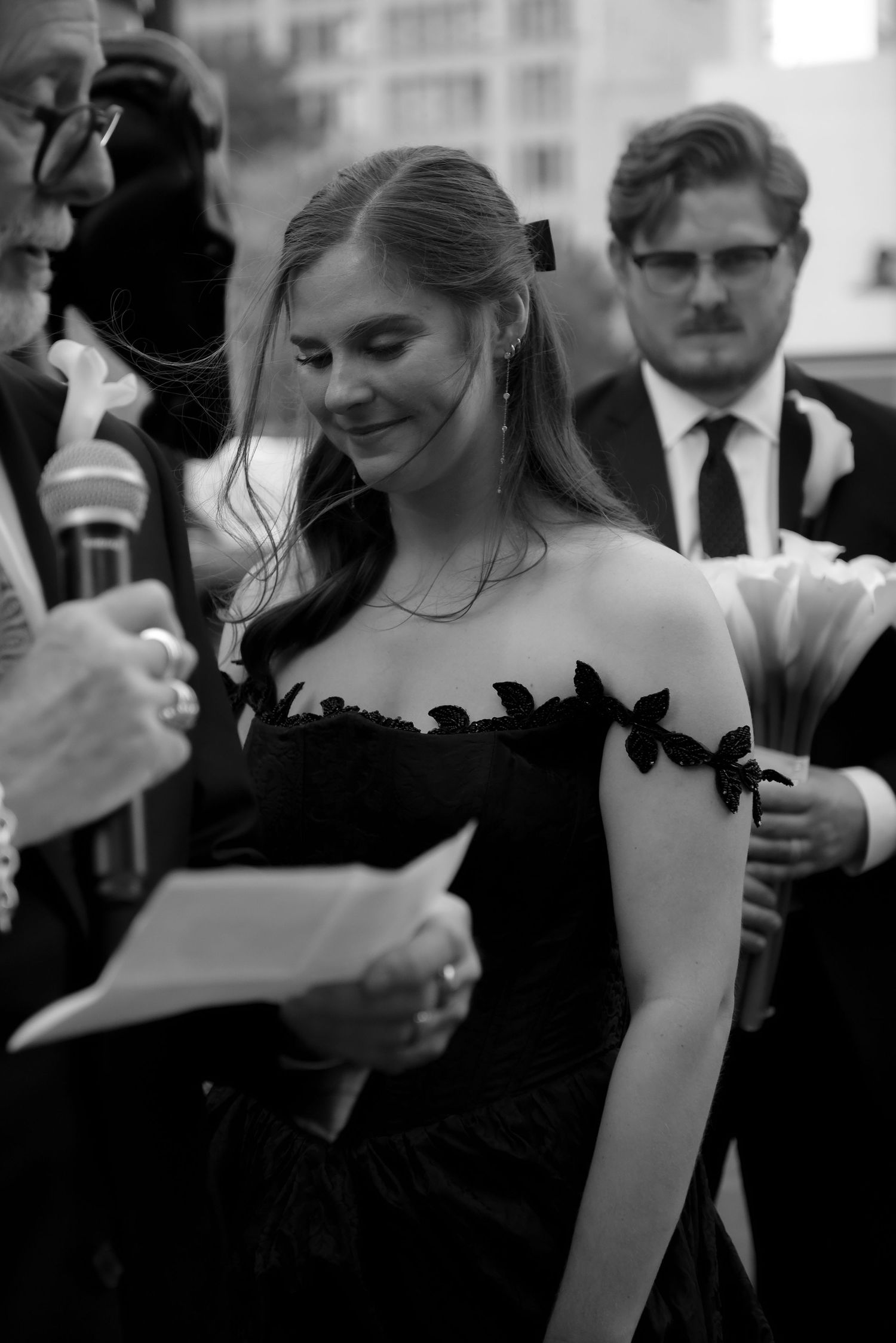 Elegant figure in a black strapless gown with floral appliques stands amidst a crowd at a formal event in black and white.