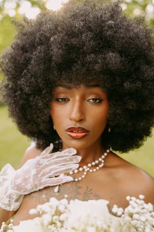 A glamorous portrait featuring an elegant afro hairstyle, pearl necklace, and white lace gloves against a soft bokeh background.