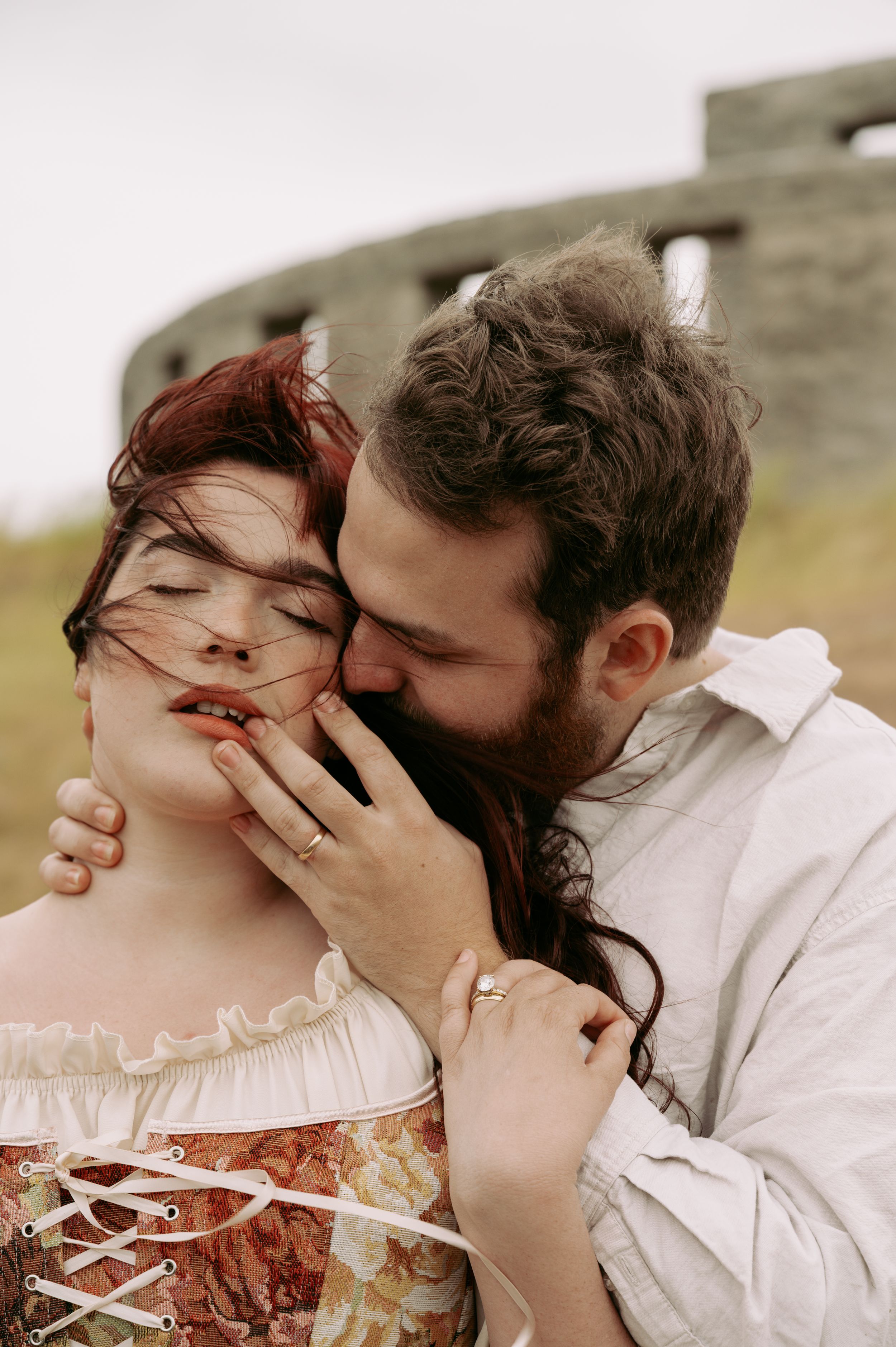 A bearded man tenderly kisses a red-haired woman in a floral corset outdoors near stone ruins.