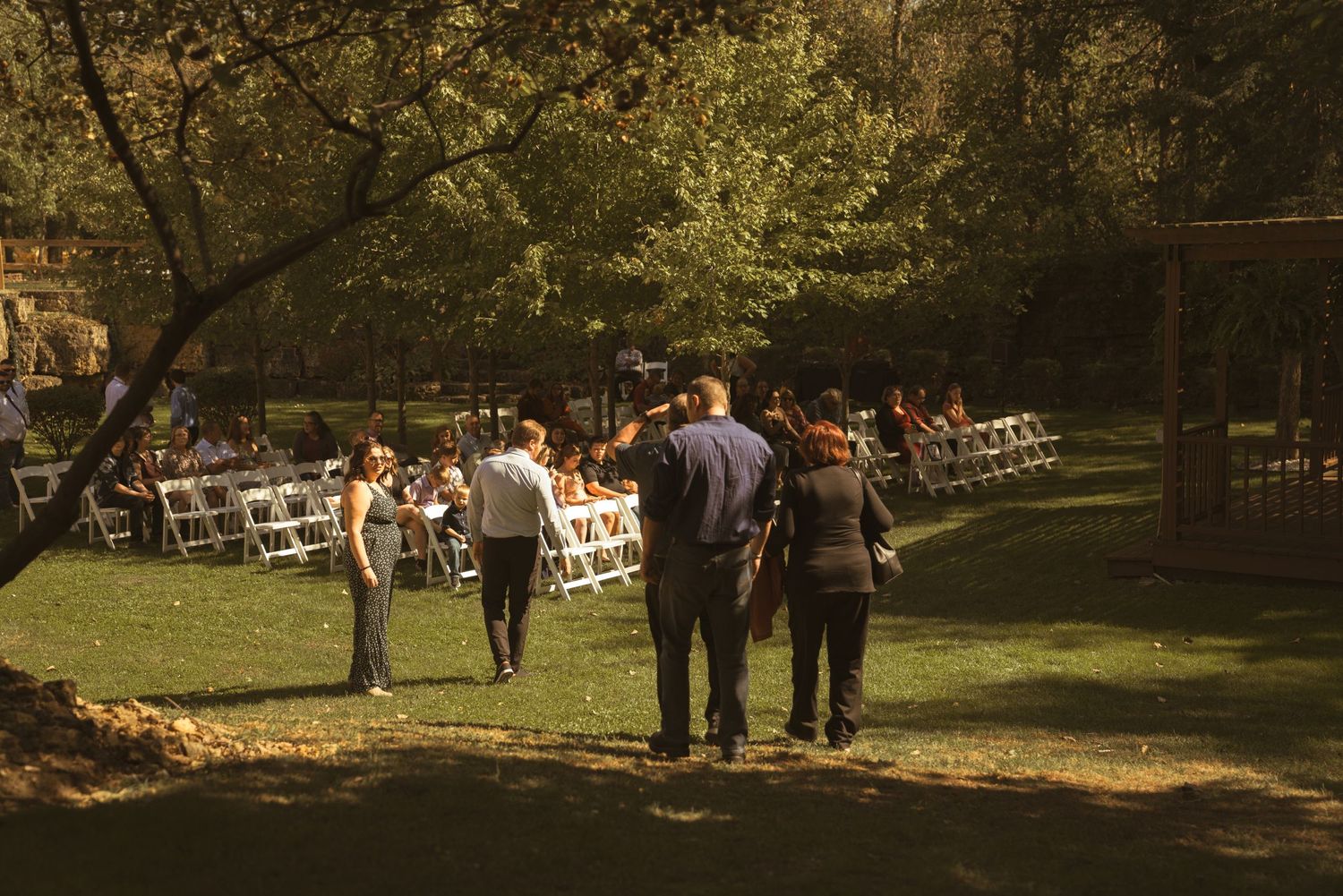 Wedding guests gather on a sunny lawn with trees and outdoor seating arrangements.