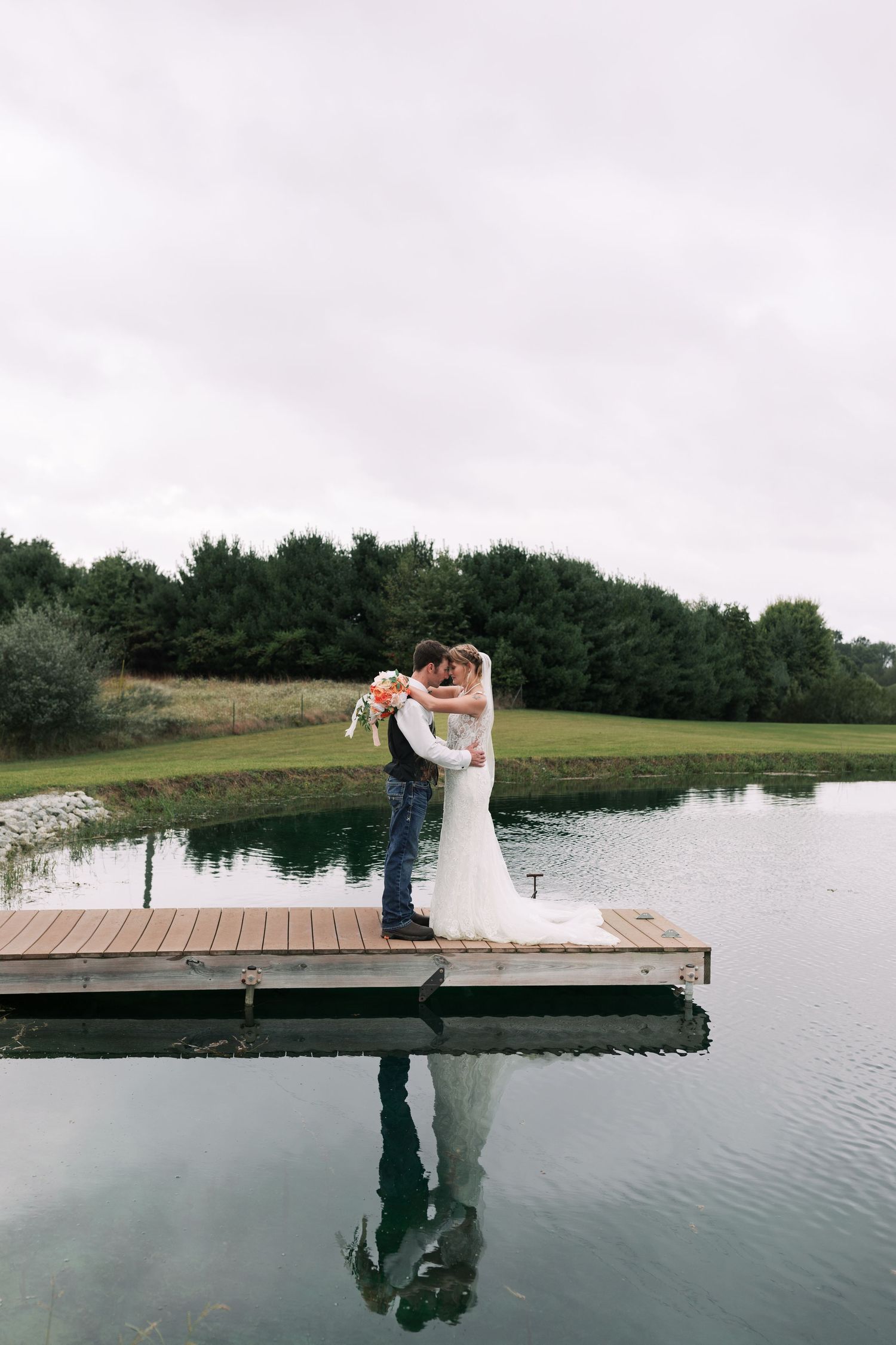 Wedding couple embracing on wooden dock over serene lake with forest backdrop.