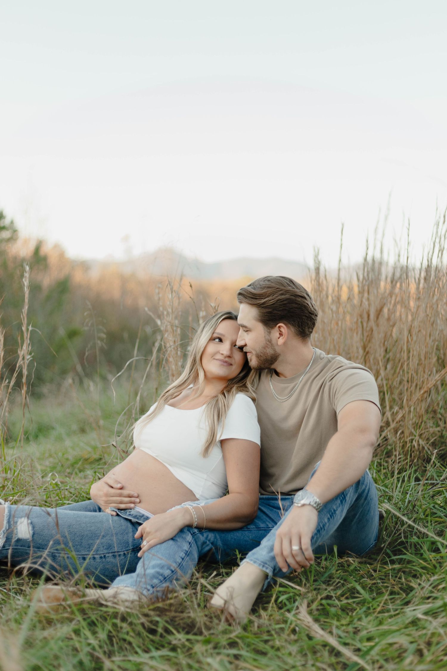 Expectant couple relaxing in tall grass during sunset maternity photo session.