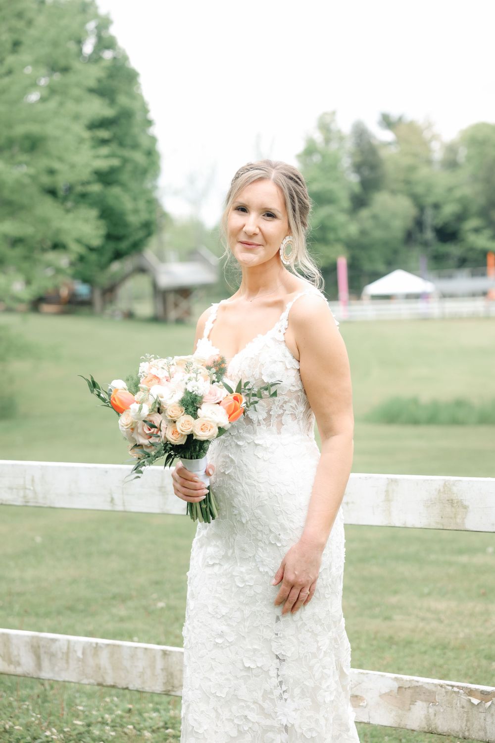 Bride holds peach and white bouquet while standing beside rustic white fence.
