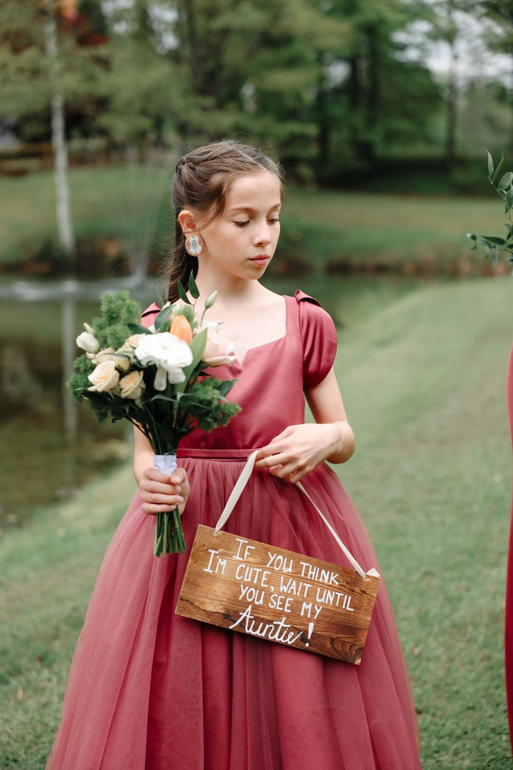 Young attendant in a rose-colored dress holds flowers and wooden sign at outdoor wedding.