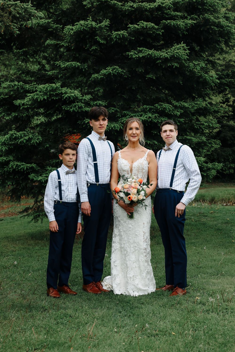 Wedding portrait with three companions in navy suspenders and white shirts against evergreen backdrop.