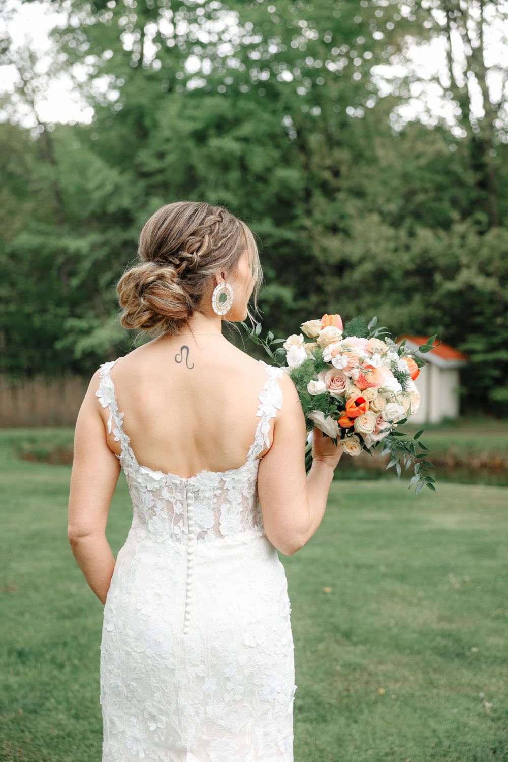 Back view of a bride in a lace dress holding a peach and white bouquet in a garden setting.